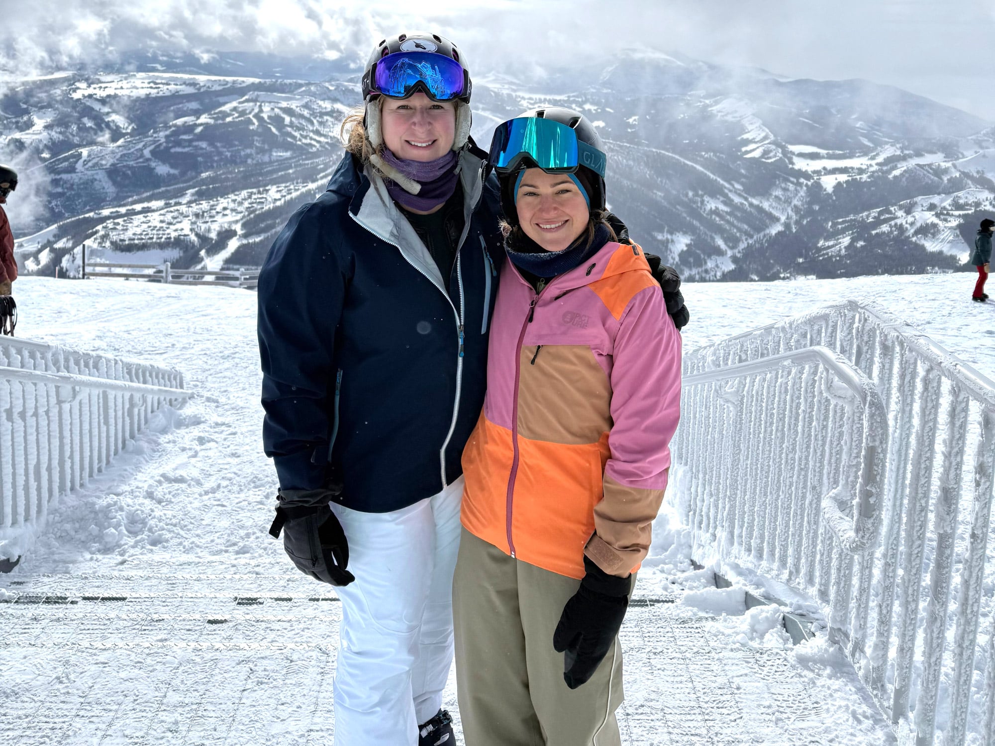 Two people in ski gear pose on snowy mountain with ski tracks and picturesque, snow-covered peaks in the background.