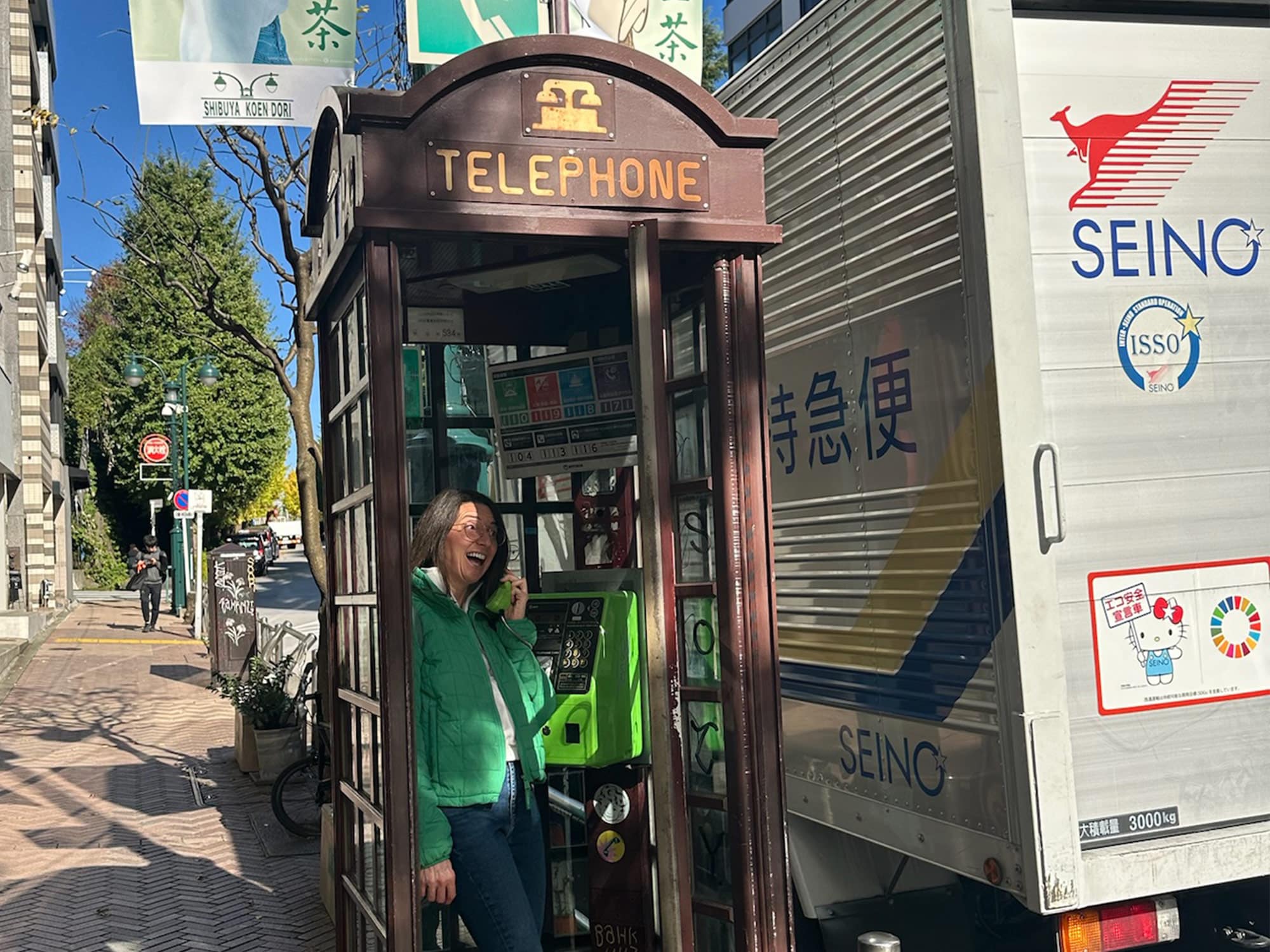 A person in a green jacket inside an old-style telephone booth on a sunlit street, near a parked delivery truck with Japanese text.