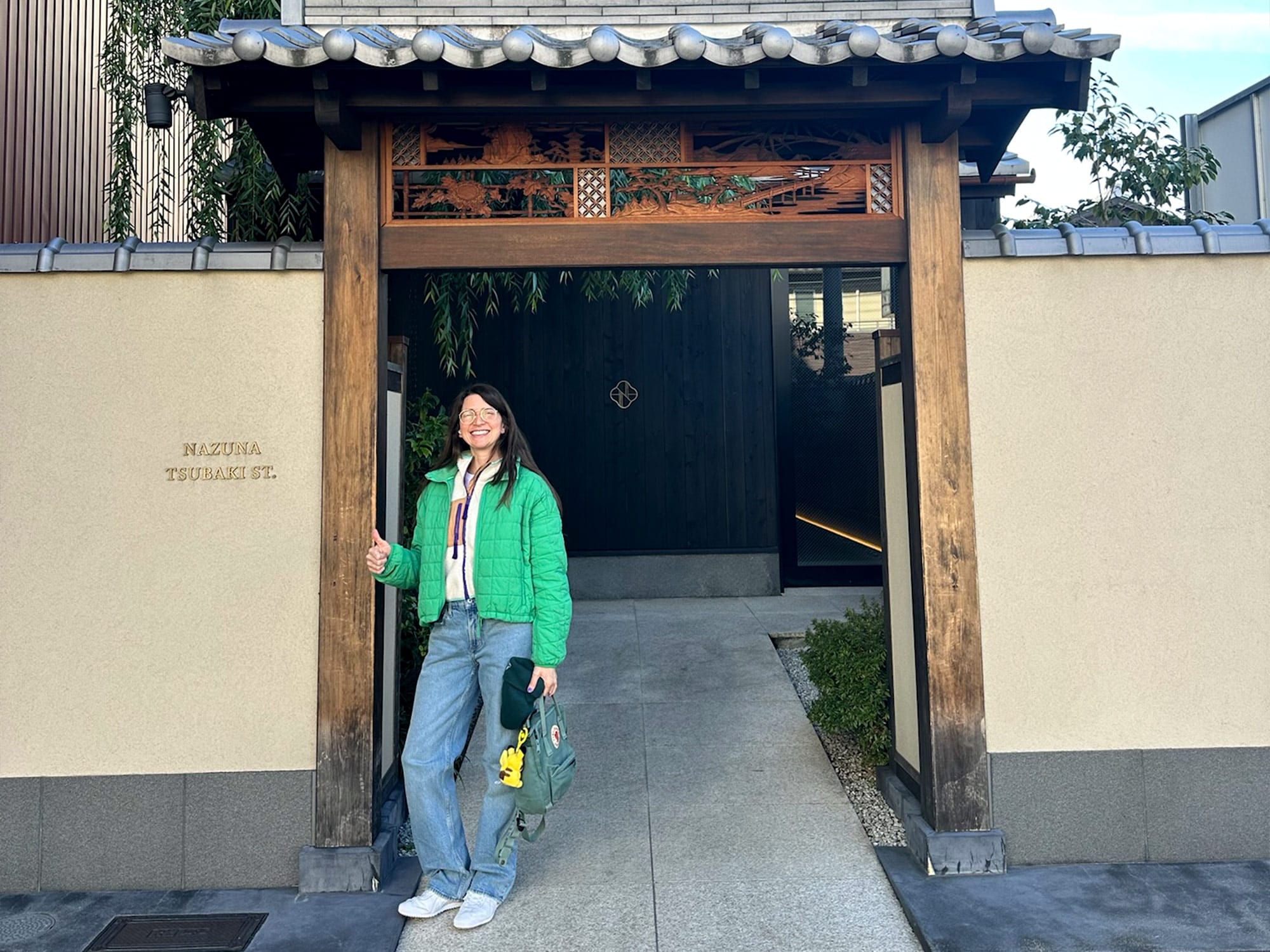 A person in a green coat stands by a traditional Japanese gate at Nazuna Tsubaki St., smiling and holding a bag.