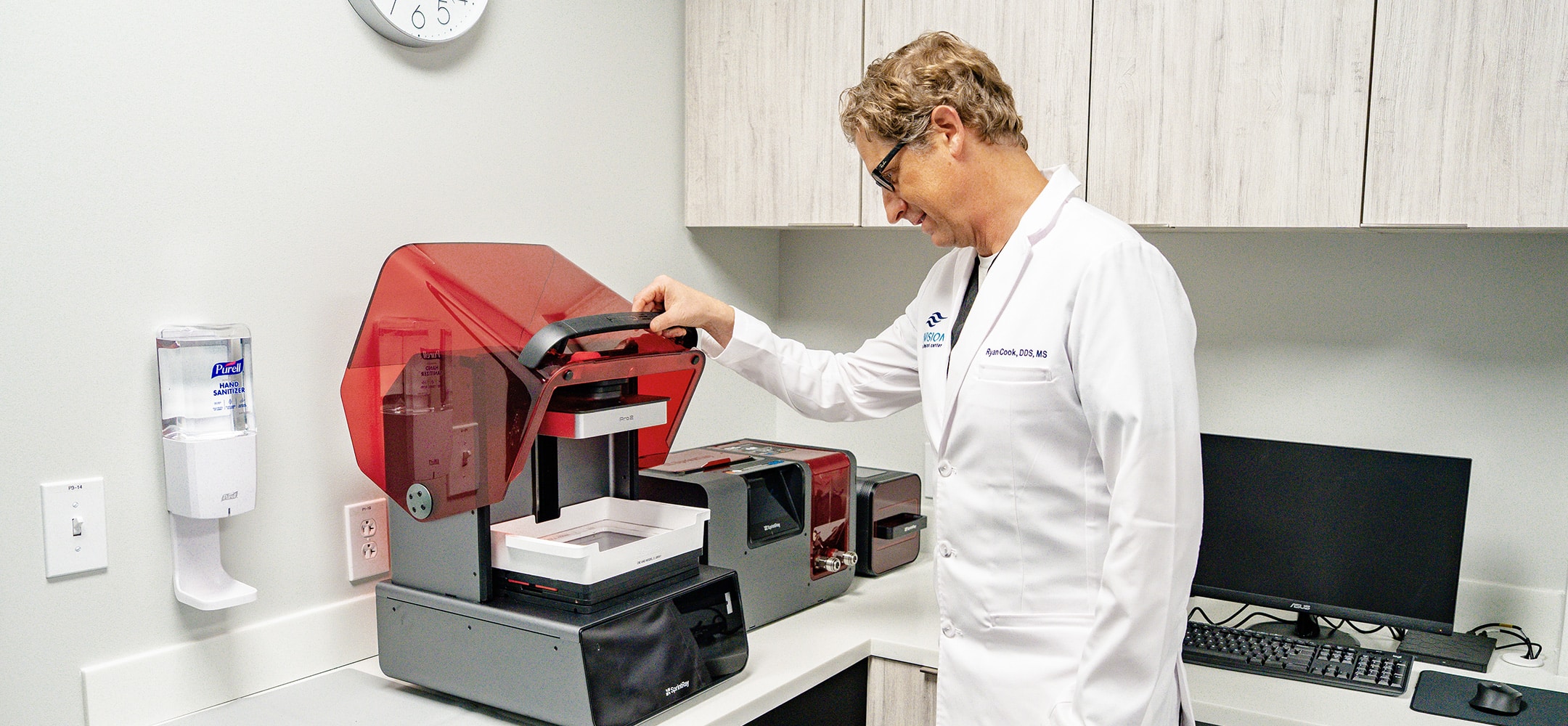 A person in a lab coat examines a 3D printer in a clinical setting, with a computer and hand sanitizer nearby.