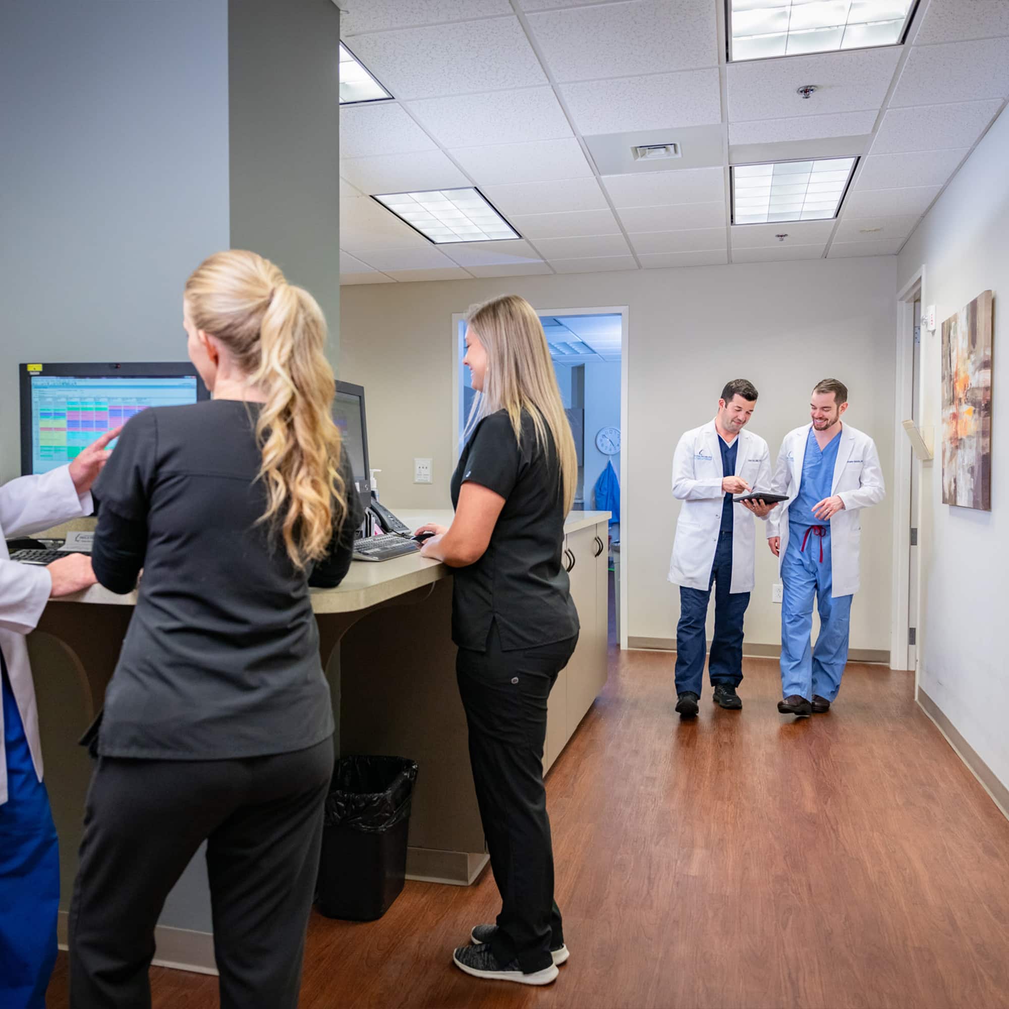 Medical office scene with four individuals in scrubs and lab coats, two standing at computers, two walking and reviewing a document.