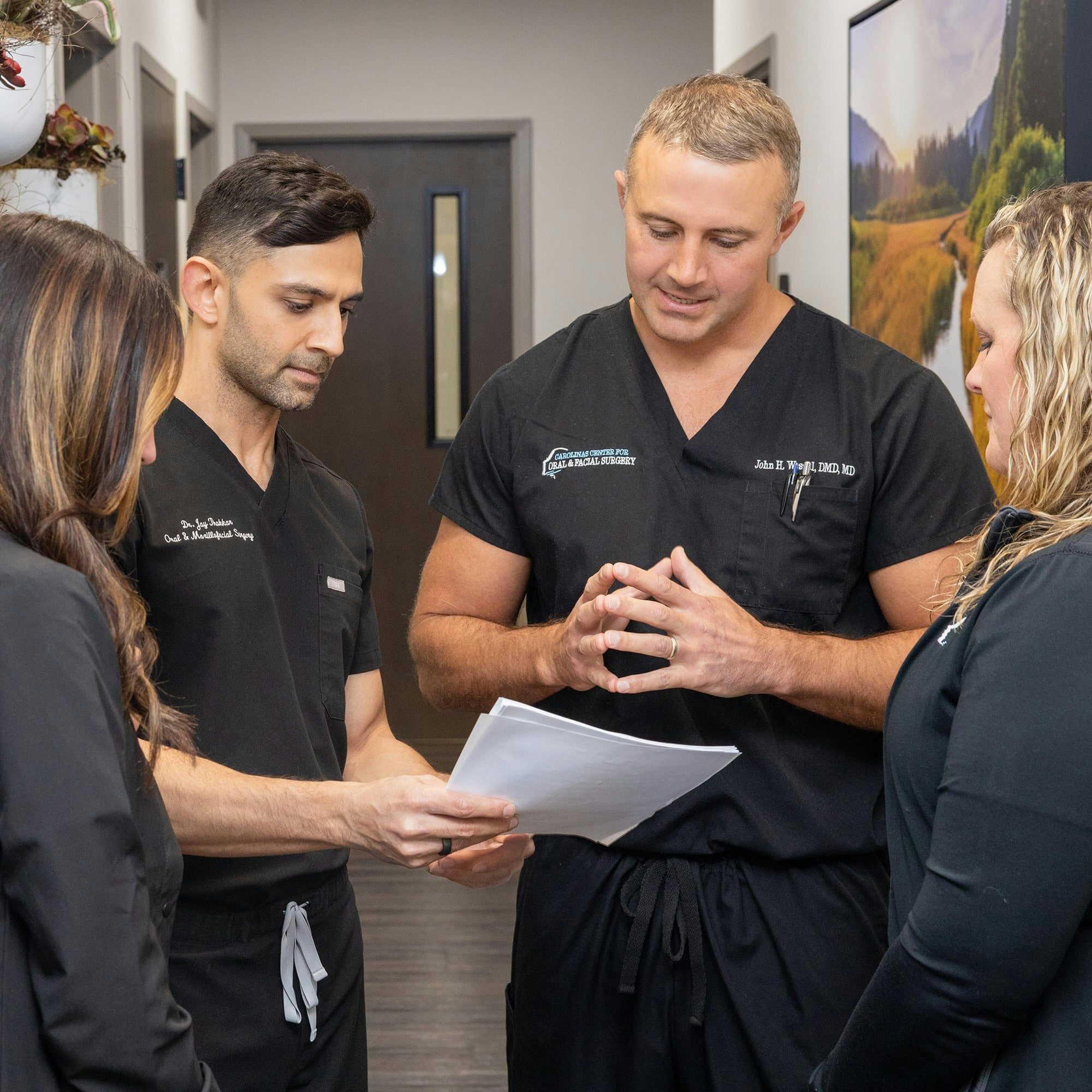 Four people in black scrubs are discussing papers in a hallway. They're likely in a medical or professional setting.