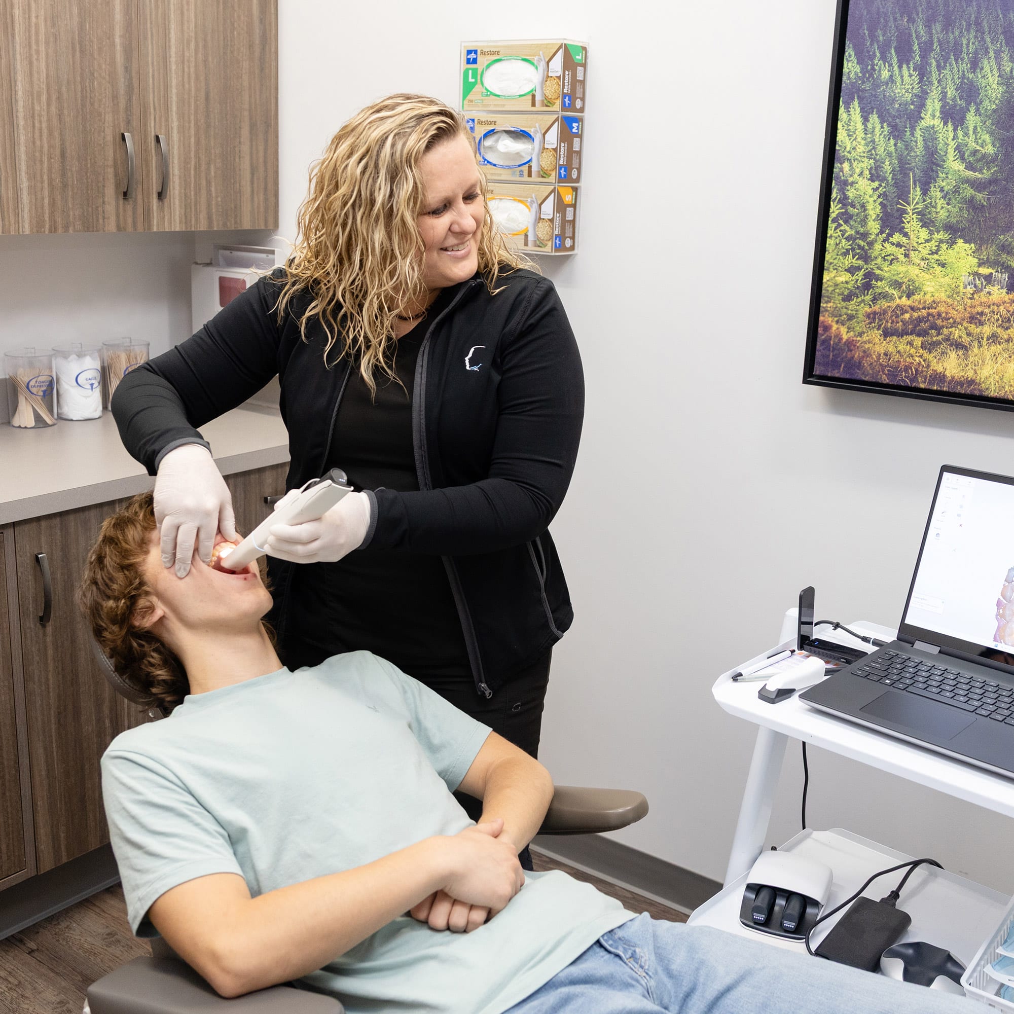 A person in scrubs examines a reclining person's mouth in a modern dental office, equipped with a laptop and dental tools.