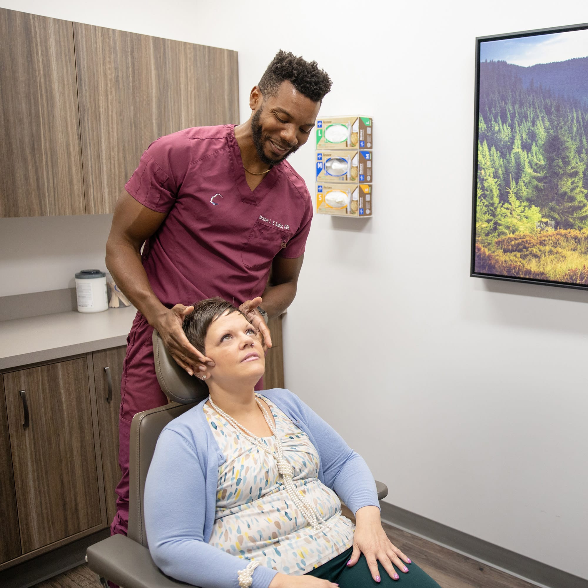A person in maroon scrubs assists another person seated in a medical office, with a forest landscape picture on the wall.