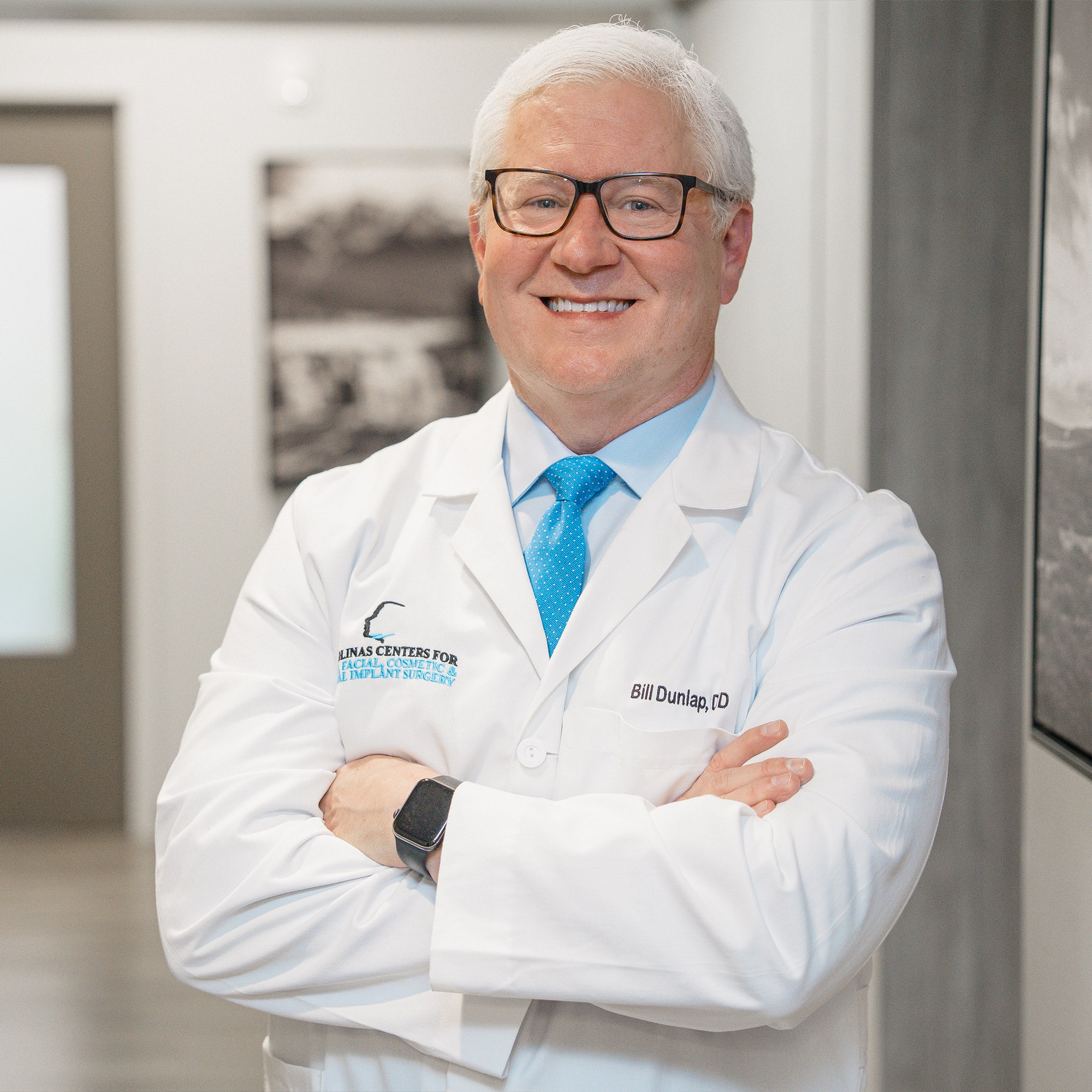 A person in a white coat poses confidently in a professional setting, with a bright tie, corridor, and framed pictures in the background.