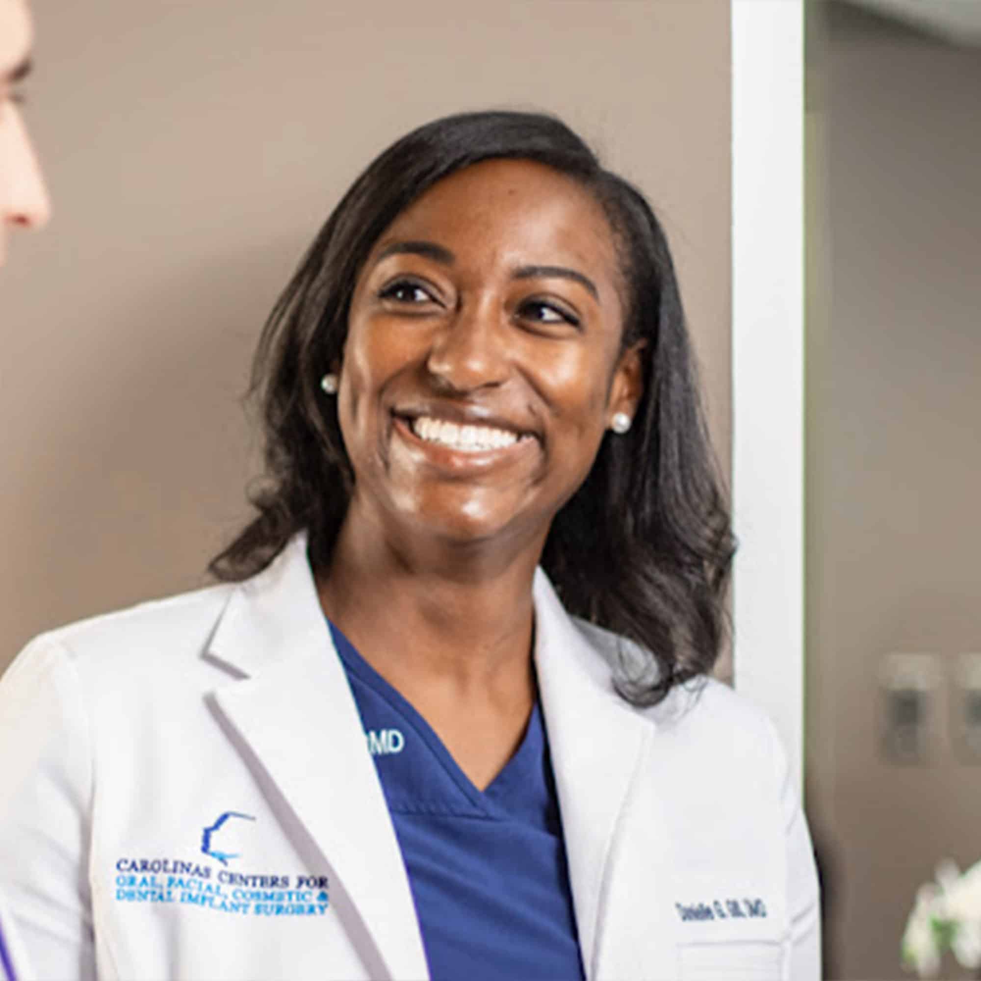 A smiling person in a white coat stands indoors, engaging with another person. They appear to be in a professional healthcare setting.