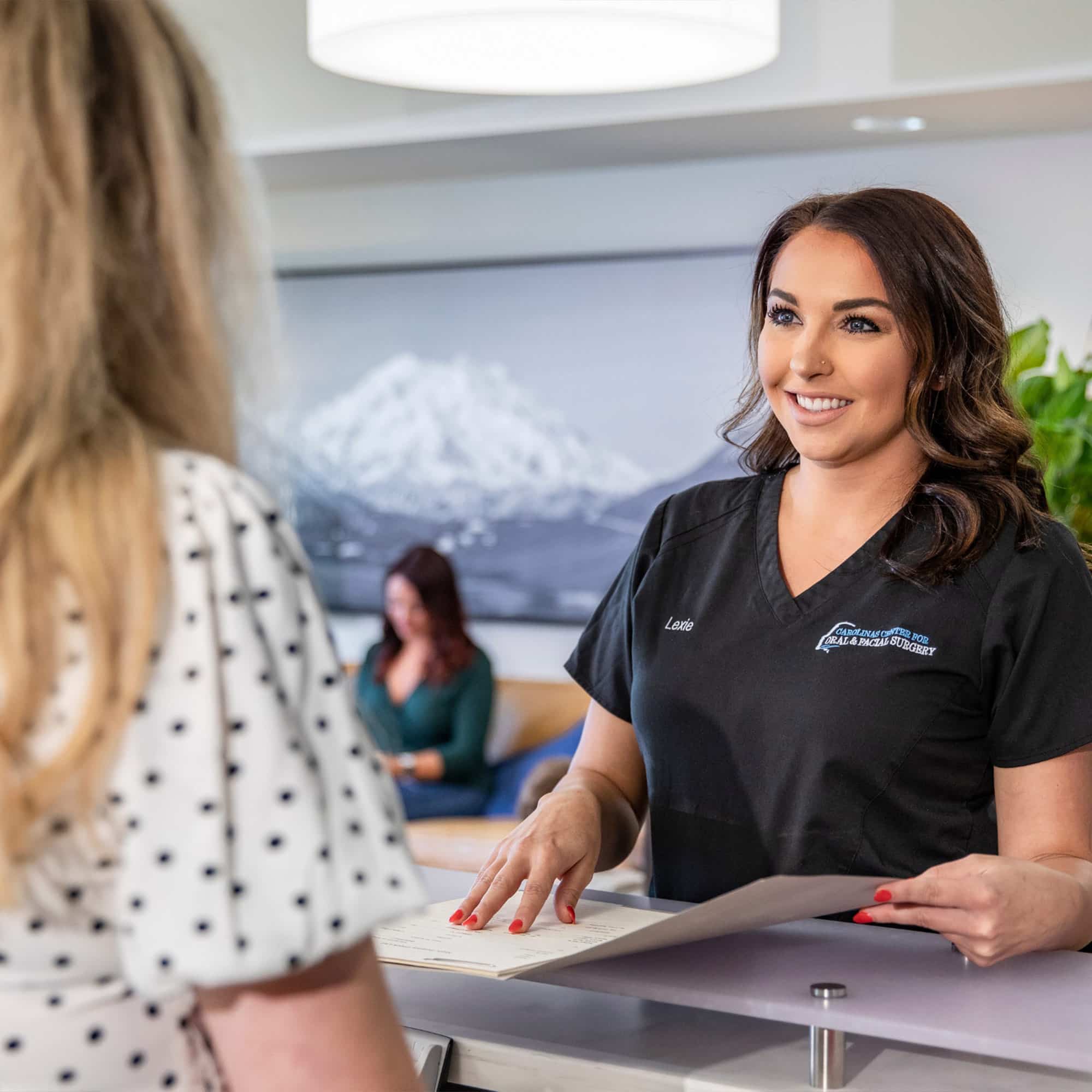 A person in black scrubs smiles at another person near a counter with paperwork. Blurred mountain photo and seated person in background.