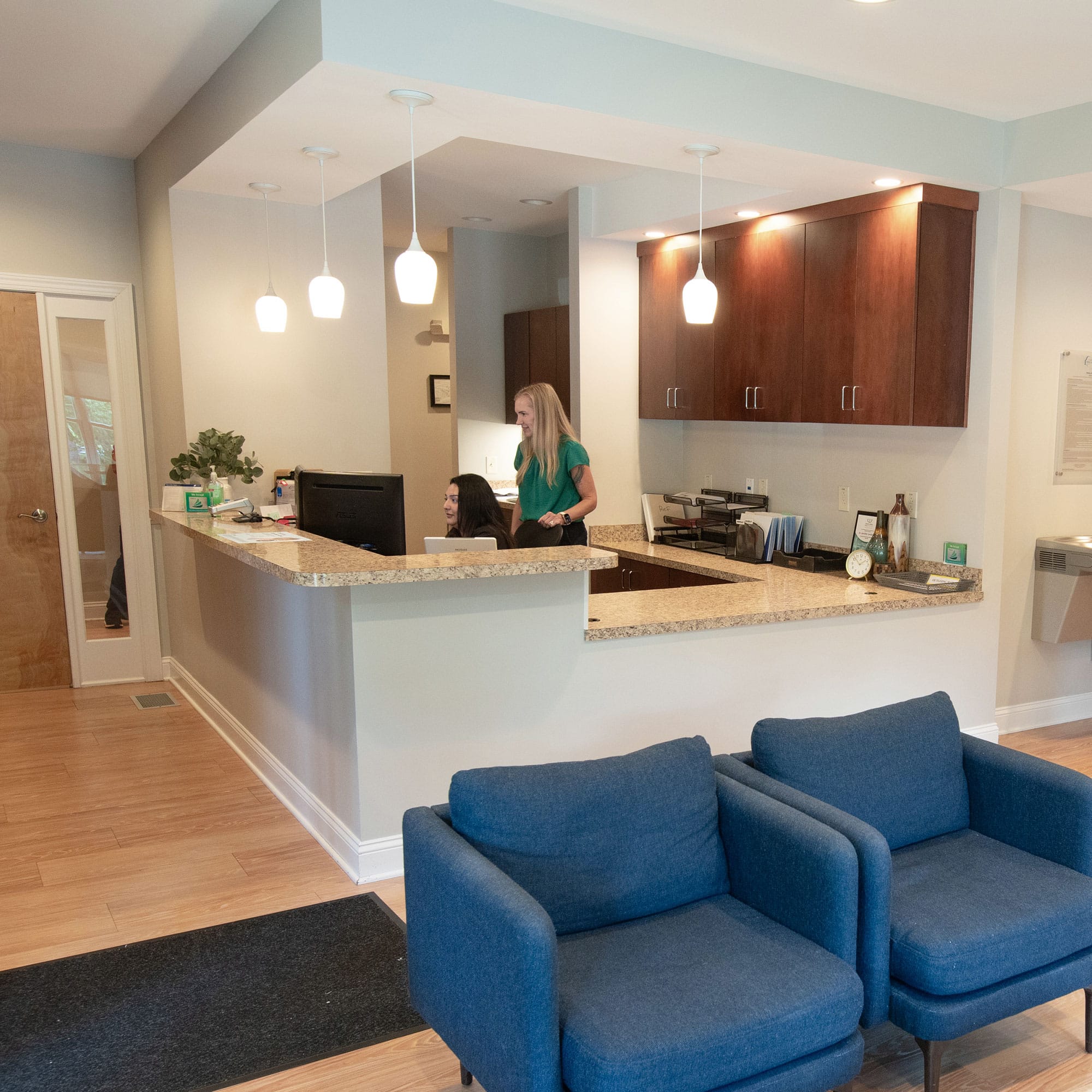 A modern, well-lit office reception area with two people at a desk, blue chairs, and wooden cabinetry.