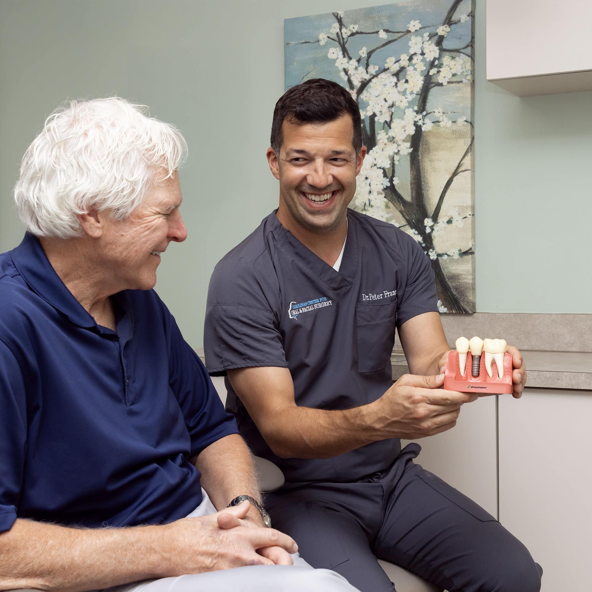 Two people smile during a dental consultation. One person holds a dental model, seated in an office with floral artwork on the wall.