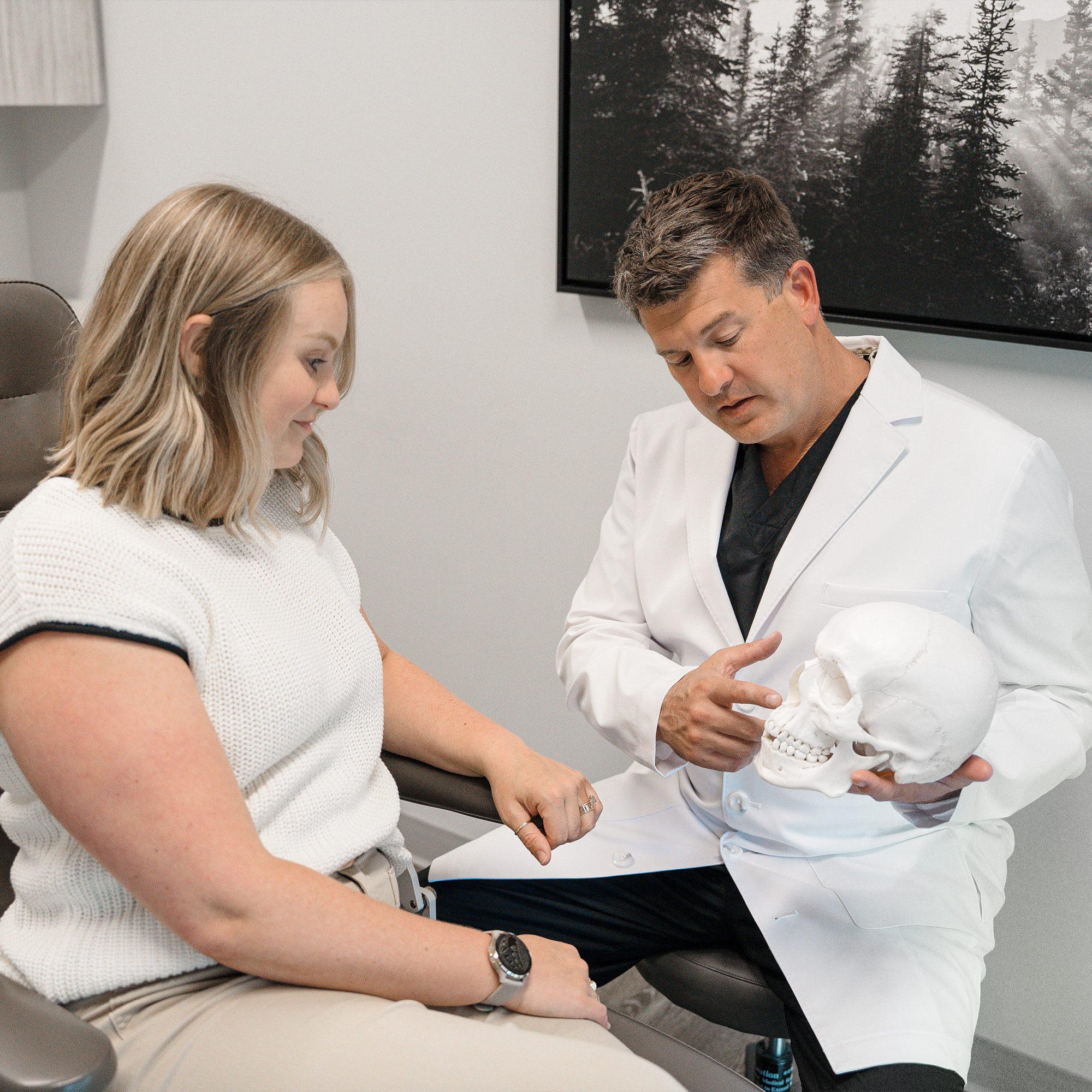 A person in a white coat discusses a skull model with another person in an office, featuring a forest photograph on the wall.