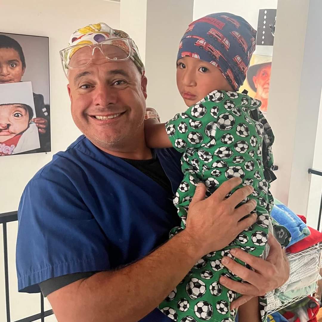 A person in blue scrubs holds a child wearing soccer-themed clothing. Colorful artwork of children is displayed in the background.
