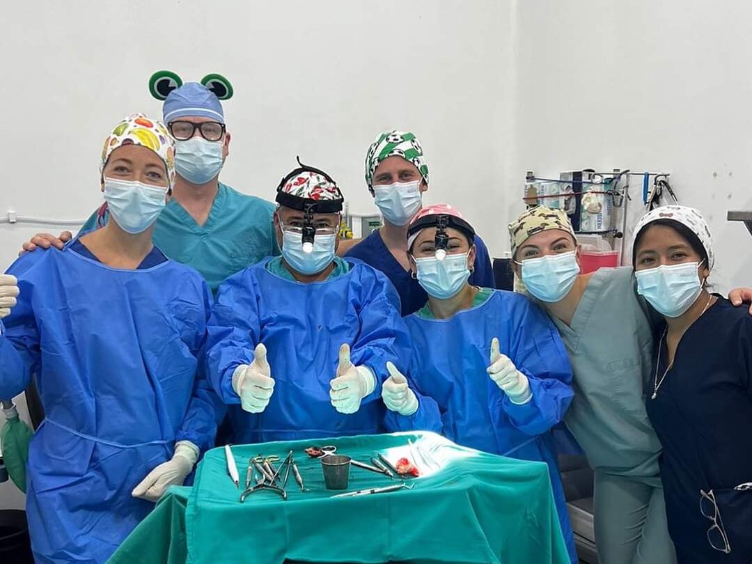 Six medical professionals in scrubs and caps pose around an operating table with surgical instruments, smiling and giving thumbs up in a hospital room.