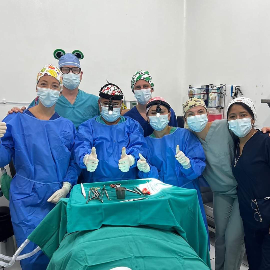 Six medical professionals in scrubs and caps pose around an operating table with surgical instruments, smiling and giving thumbs up in a hospital room.