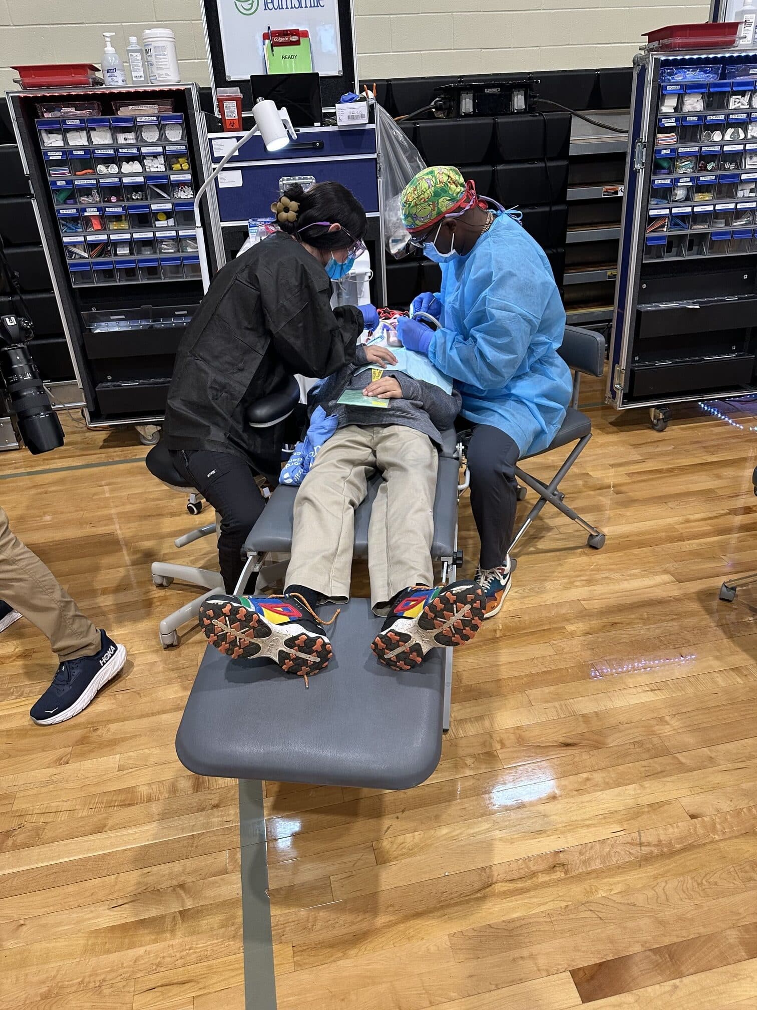 Two people provide dental care to a seated person in a clinic setting with medical supplies on a hardwood floor.