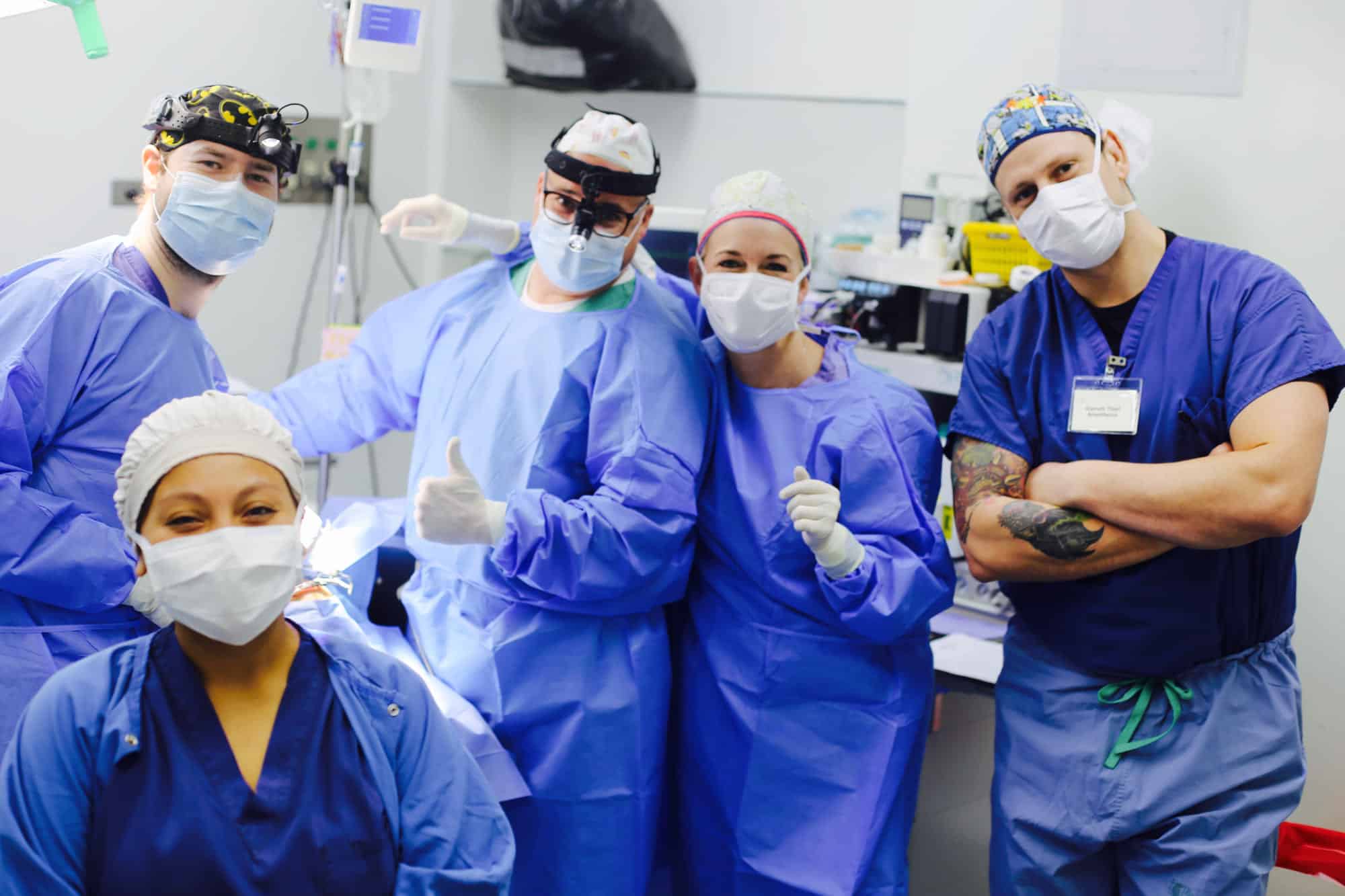 Five medical professionals in surgical attire pose together, smiling and giving thumbs up, in a brightly lit hospital room.