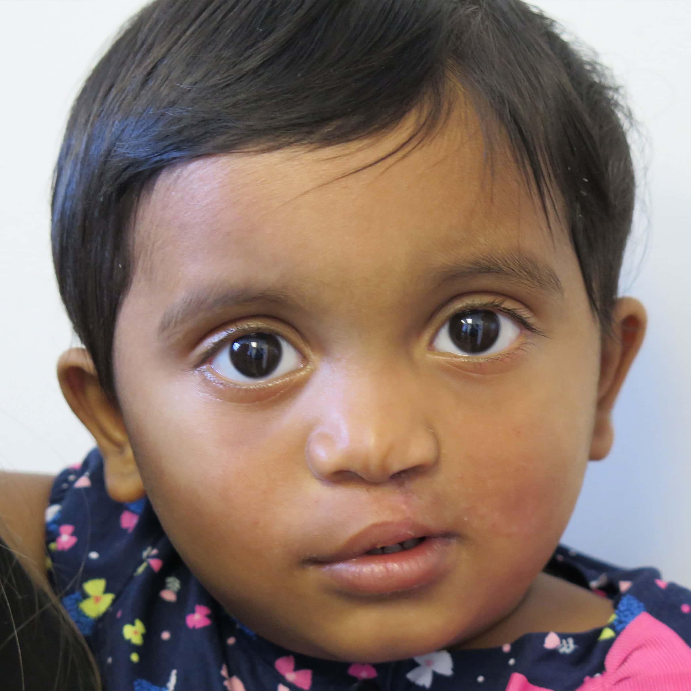 A close-up of a young child with short dark hair, wearing a floral-patterned outfit, and looking directly at the camera.