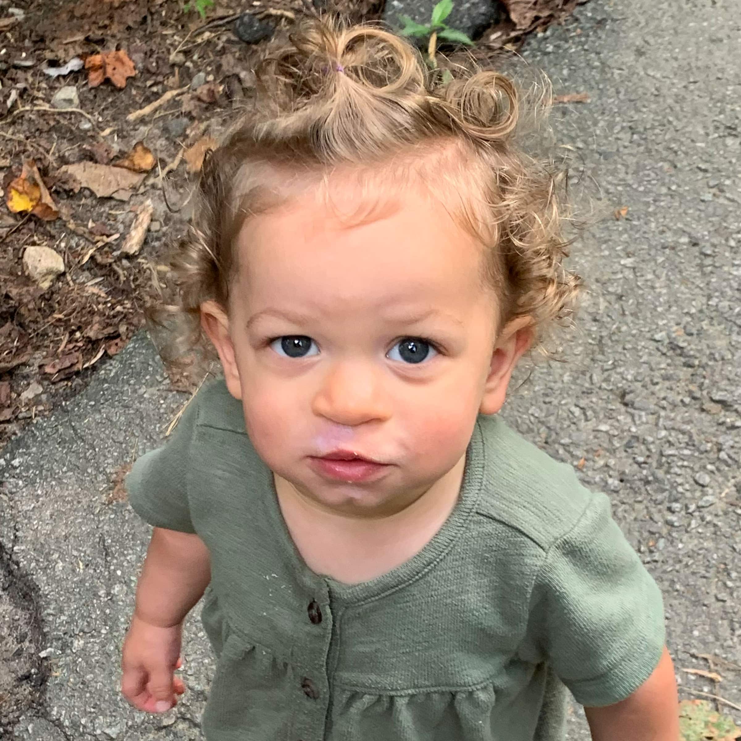 A toddler with curly hair wearing a green shirt stands on a rocky path, looking up with curious eyes, outdoors surrounded by foliage.
