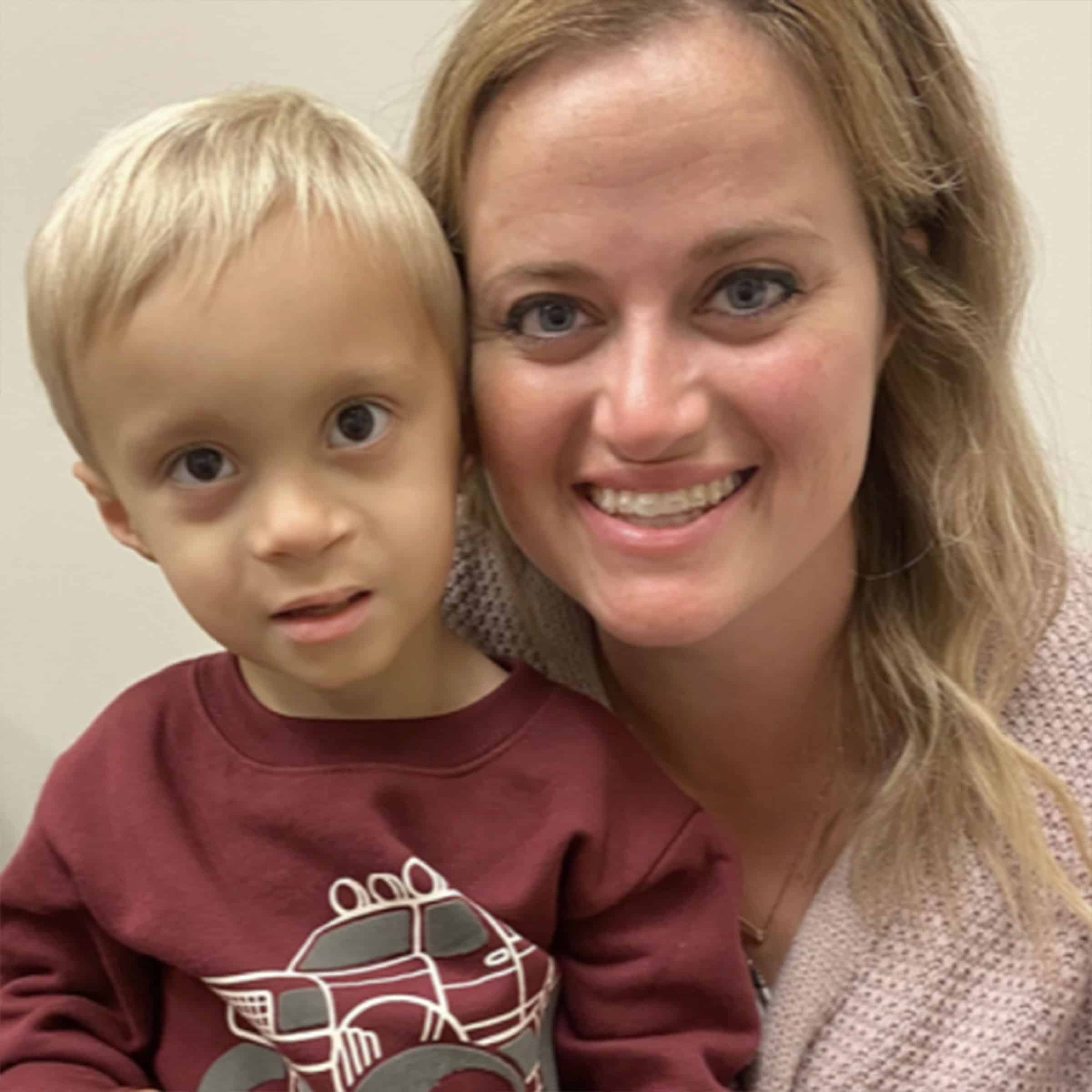 A smiling person and child pose closely together indoors, with the child wearing a sweatshirt featuring a vehicle design.