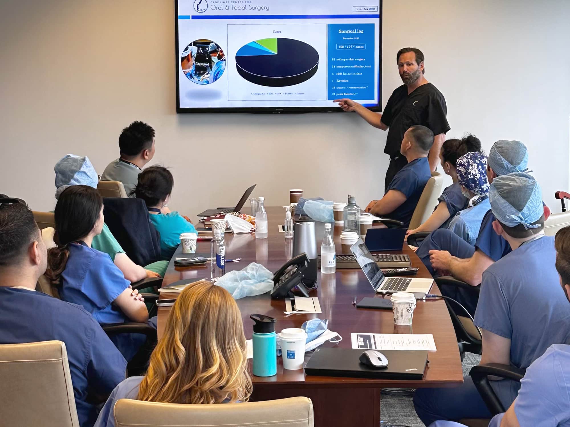 A person presents oral surgery data to a group in medical attire, seated around a conference table with laptops and charts.