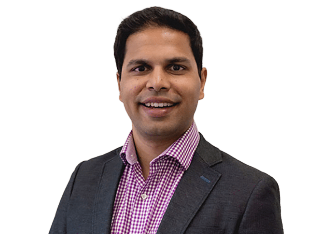 A person in a checkered shirt and blazer smiles confidently against a plain background, conveying a professional and approachable demeanor.