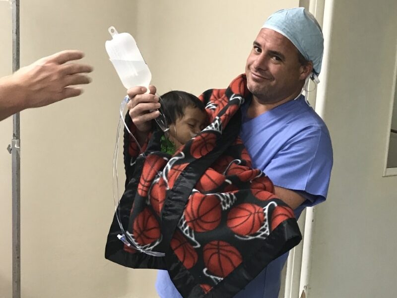 A person in scrubs holds a child wrapped in a basketball-patterned blanket, carrying an IV drip, in a hospital setting.