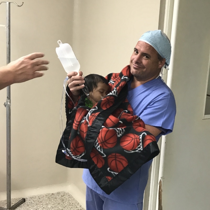 A person in scrubs holds a child wrapped in a basketball-patterned blanket, carrying an IV drip, in a hospital setting.