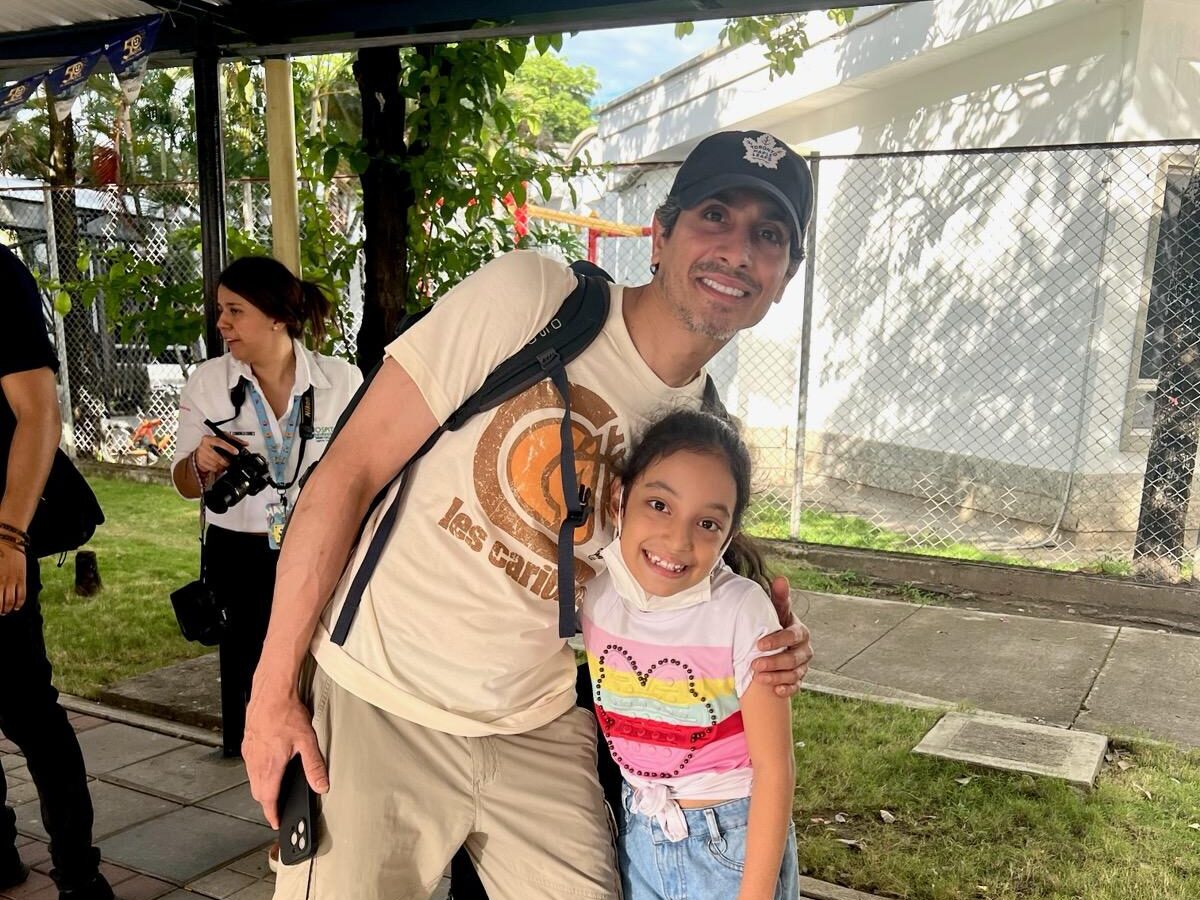A person and child smile under a metal canopy. Another person in white stands nearby. There's greenery and a chain-link fence in the background.