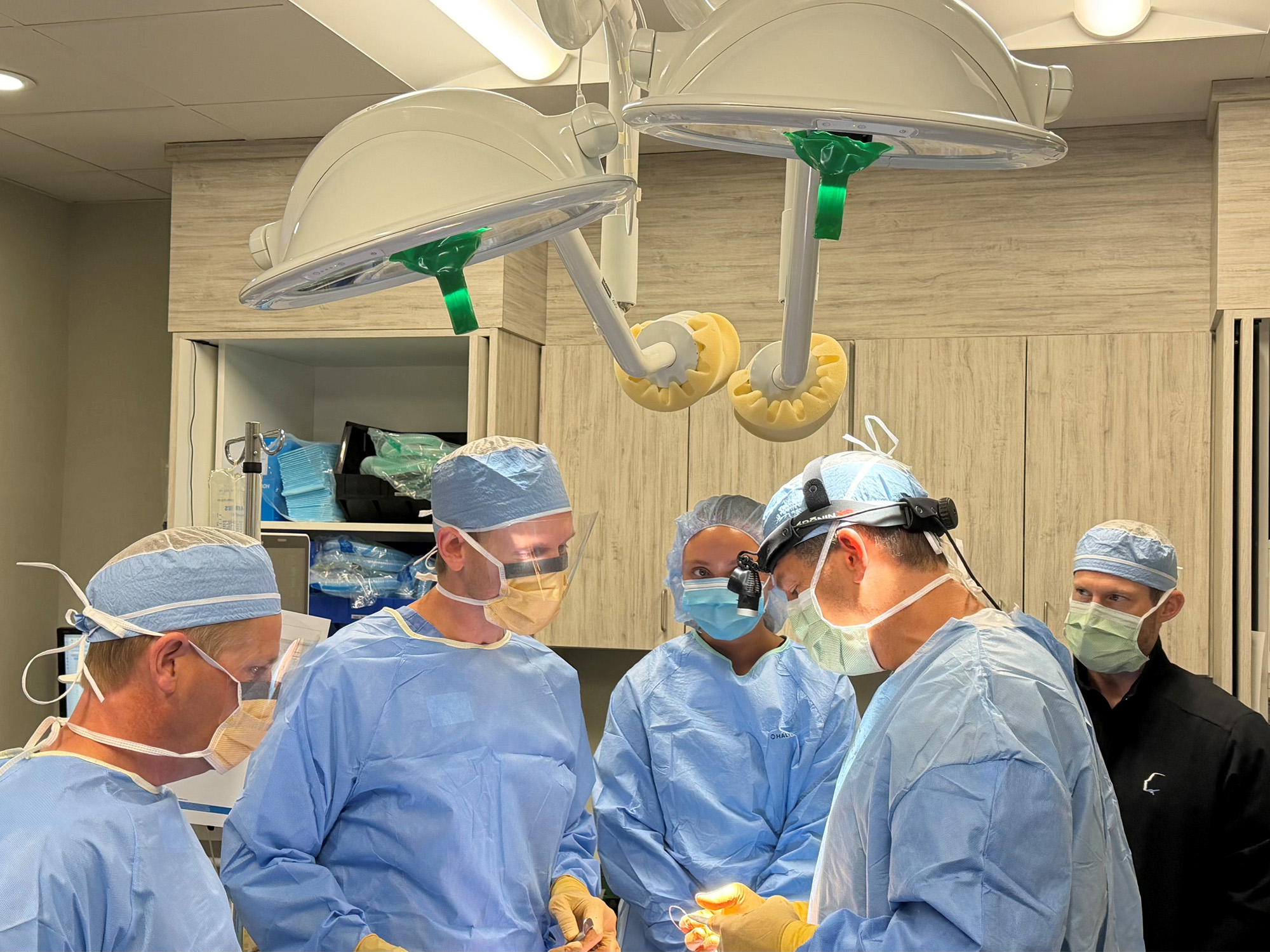 A team of oral surgeons in medical scrubs performing surgery under an operating room light. Surgical tools and IV equipment are visible in a clinical setting.