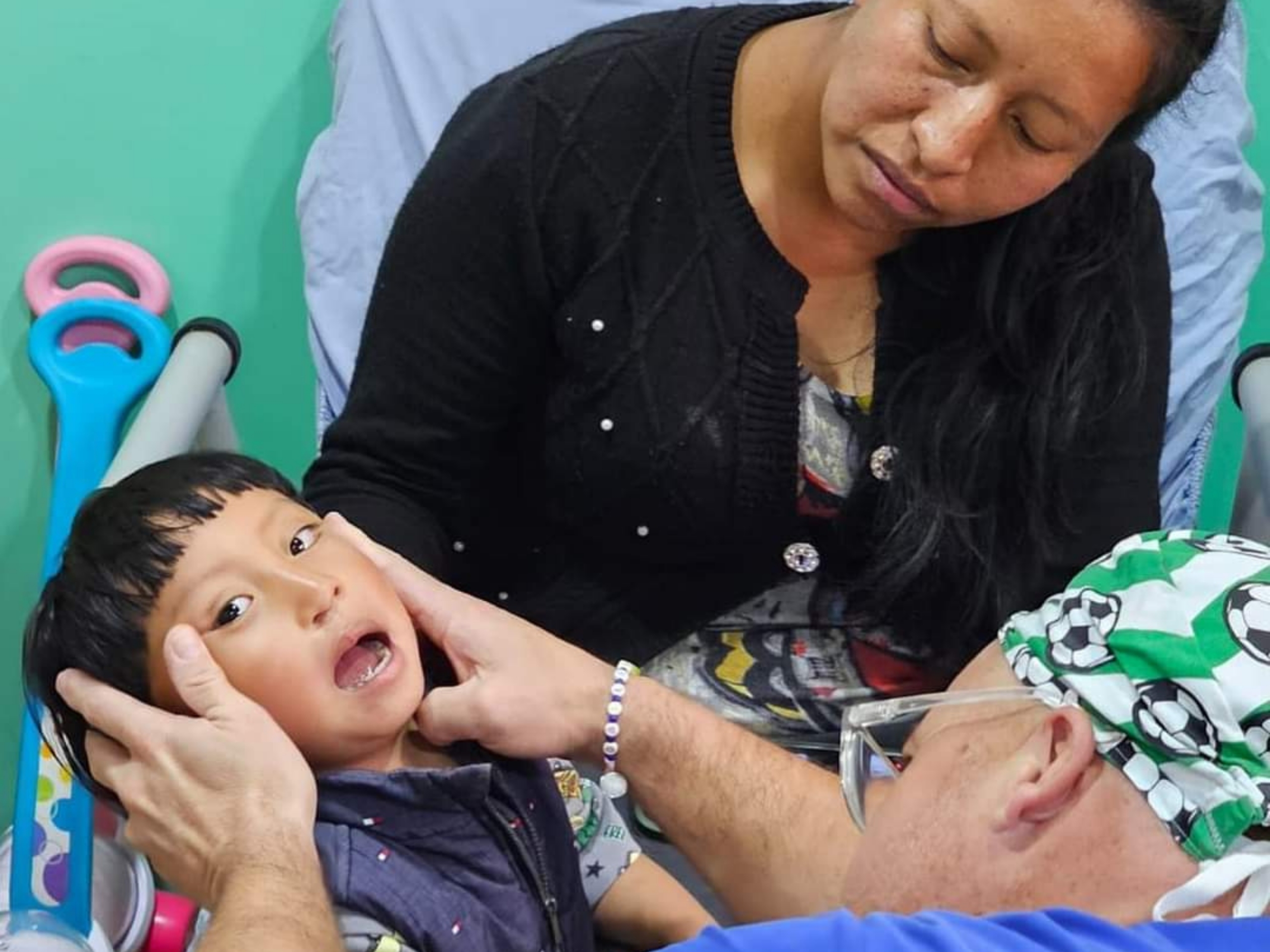 A person examines a child's mouth, while another person observes in a room with colorful toys in the background.